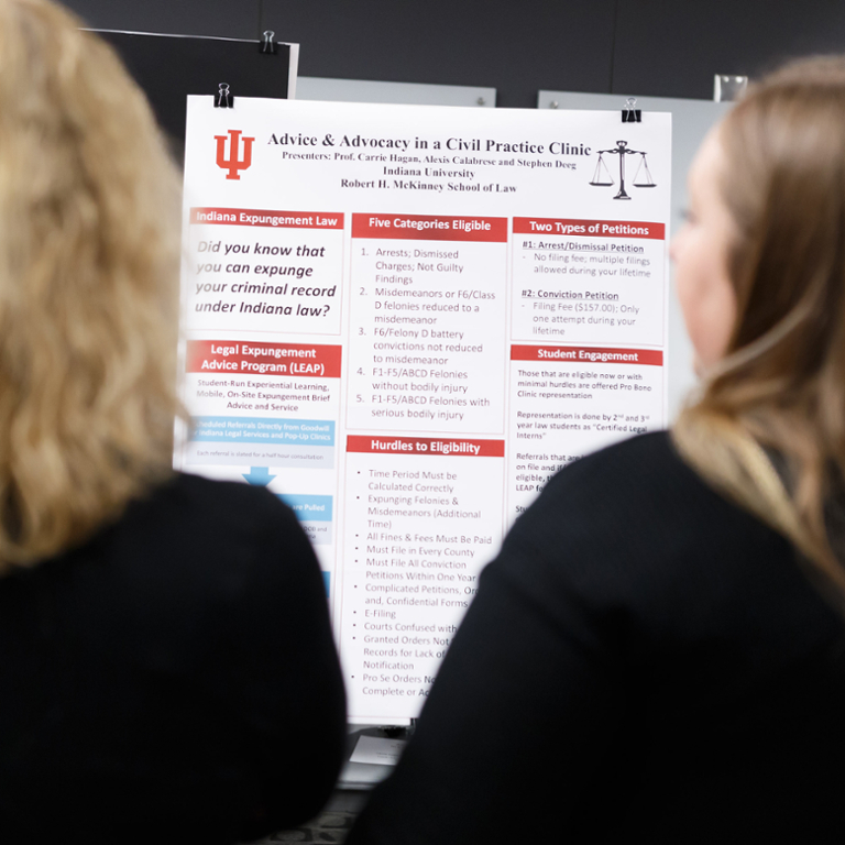 Two women standing in front of a poster on a table. The focus of the photo is on the poster in the middle.