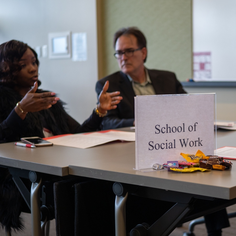 Woman at table talking to two other people, one who is not pictured, with a sign that says School of Social Work